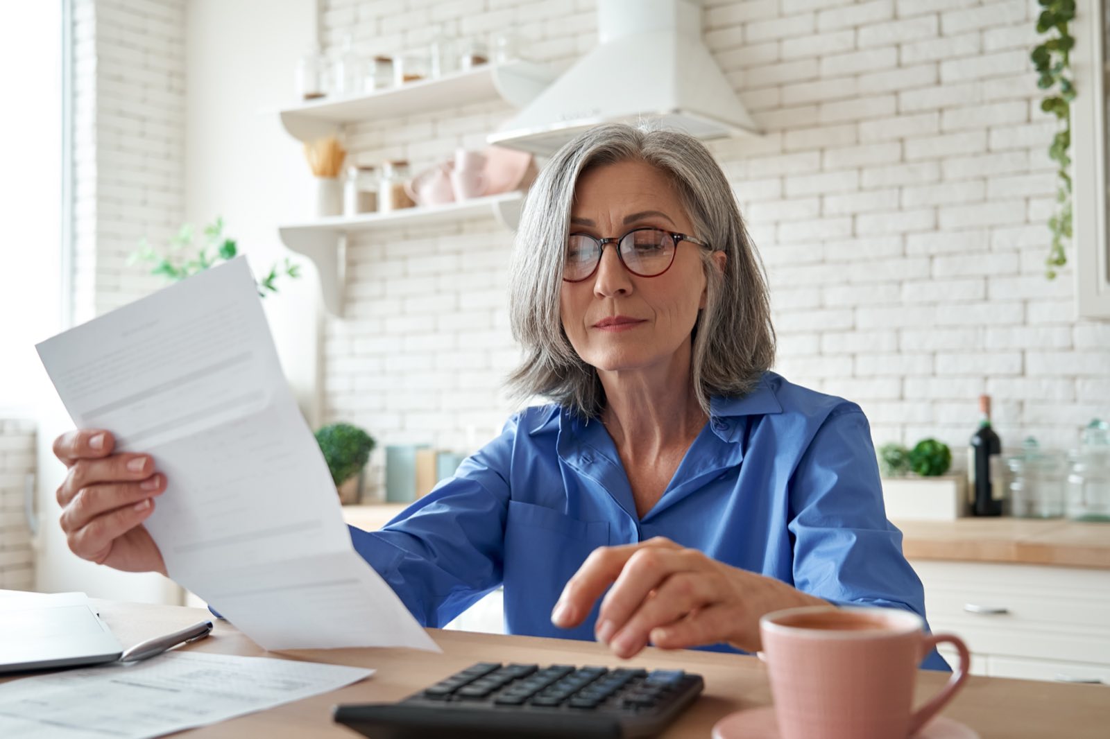 A retired woman reading a letter at her kitchen table, with a calculator